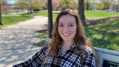 A woman in a plaid shirt and long brown hair sits on a bench outside.