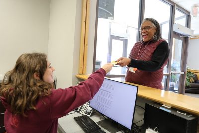 A student working at the Housing Services front desk hands a document to another person who is standing on the opposite side of the front desk. A lobby with a couch and plants can be seen behind the guest. Both are wearing Hokie colors, a maroon sweatshirt and a maroon vest respectively, and smiling. 
