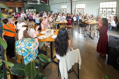 A woman speaks into a microphone as a large seated audience applauds during an indoor event.